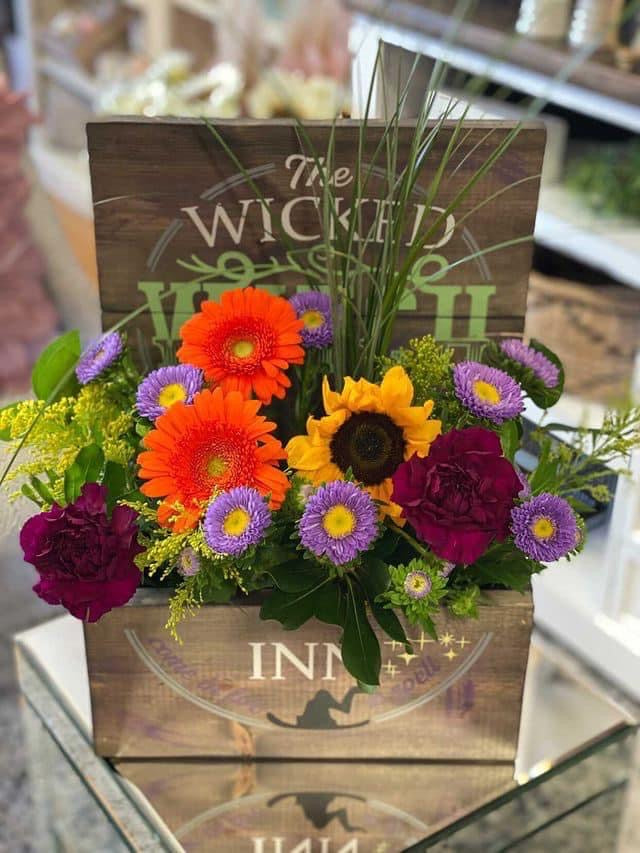 A vibrant flower arrangement in a rustic wooden box with the words 'The Wicked Inn' printed on it. The bouquet features bright orange gerbera daisies, yellow sunflowers, purple asters, and deep magenta carnations with green foliage, set on a reflective glass table.