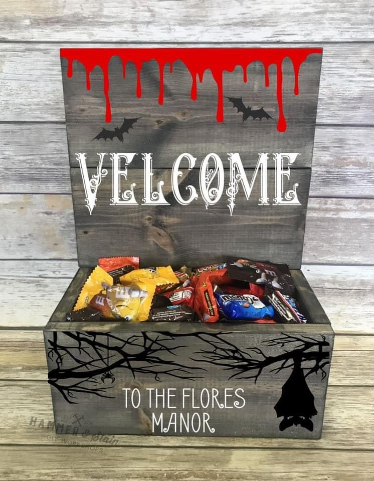 A Halloween-themed wooden gift box filled with assorted candy, featuring a rustic gray wood finish. The lid has a red dripping blood design, black bats, and the word 'WELCOME' in white.