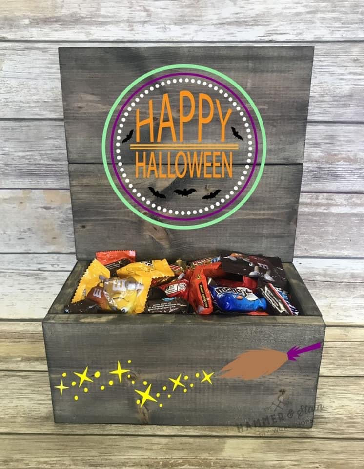 A rustic wooden Halloween gift box filled with assorted candy, featuring a painted 'Happy Halloween' sign on the lid and a broomstick graphic on the front, set against a weathered wood plank background.