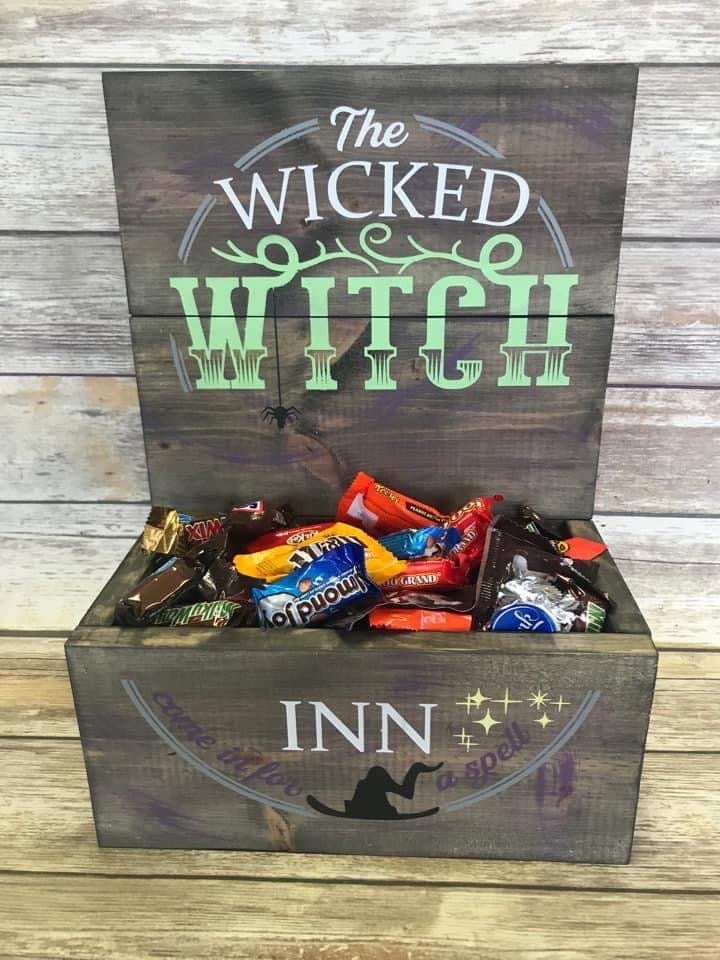 A rustic Halloween-themed wooden gift box with a hinged lid, featuring the text 'The Wicked Witch' on the lid and 'INN' with a witch silhouette on the front. The box is filled with assorted candy and is set against a gray wood-plank background.