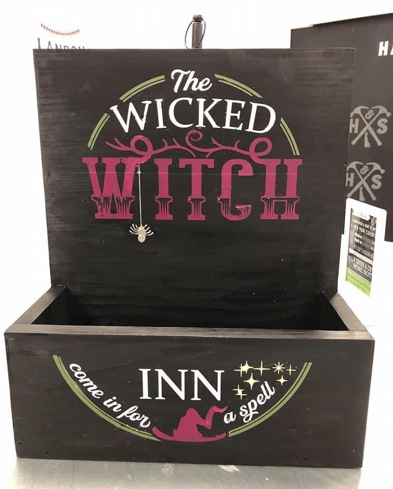 A black wooden decorative box with a hinged lid, featuring the words 'The Wicked Witch' in white and pink lettering on the lid, and 'INN come in for a spell' with a witch silhouette and stars on the front.