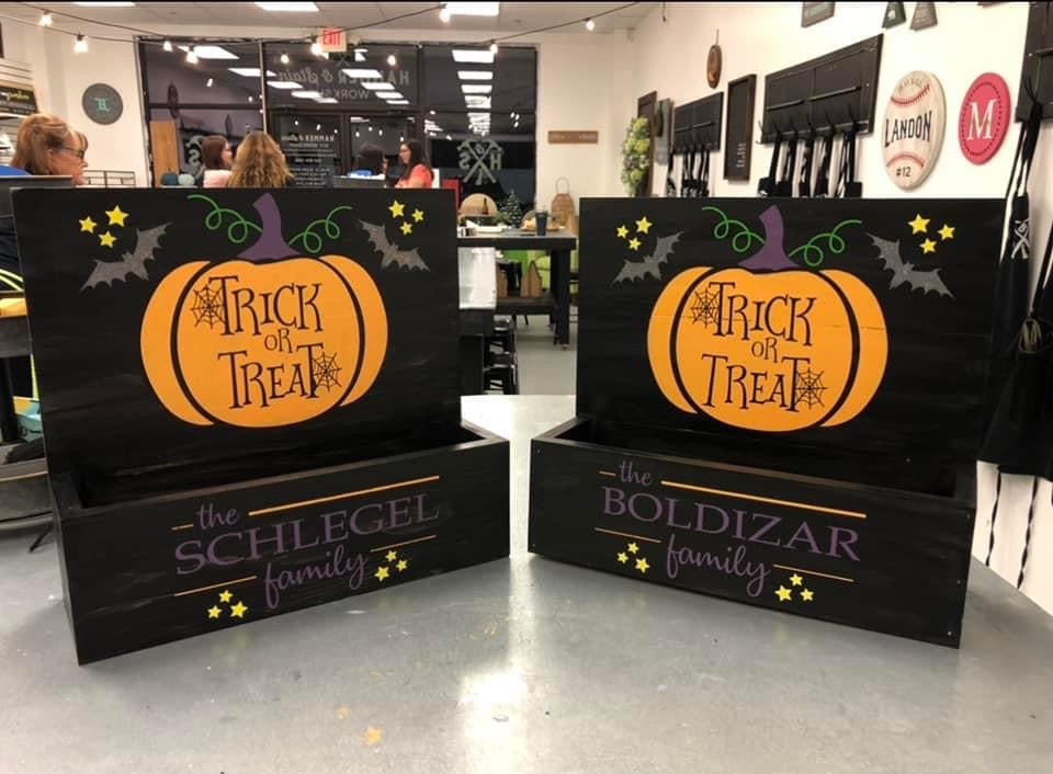 Two black wooden Halloween-themed decorative signs or planters displayed on a counter in a retail store. Each sign features a large orange pumpkin with 'Trick or Treat' written inside, along with bats, stars, and purple text reading 'the SCHLEGEL family' and 'the BOLDIZAR family'.