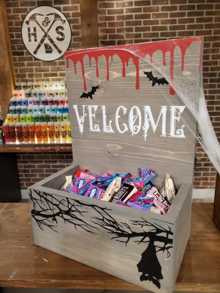 A rustic Halloween-themed wooden candy box with a hinged lid, decorated with 'VELCOME' in white, red dripping paint, black bats, and black tree branch silhouettes. The box is filled with assorted candy and sits on a wooden counter in front of a brick wall.