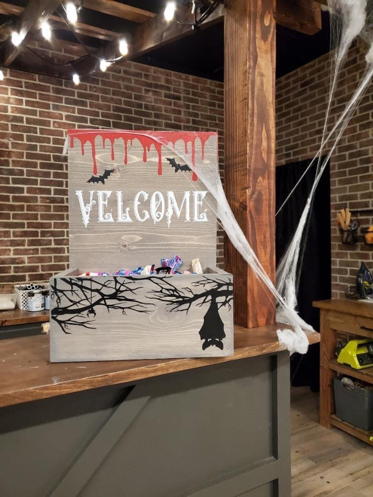 A rustic Halloween-themed wooden welcome box with a painted sign reading 'WELCOME' in white, red dripping paint at the top, black bats, and a black bat silhouette on the front. The box sits on a wooden counter in a decorated room with brick walls, hanging webbing, and string lights.