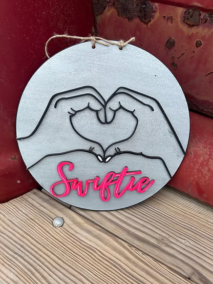 Decorative plaque with hands forming a heart and 'Swiftie' text on a rustic background