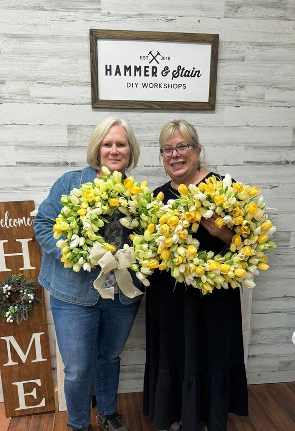 Two women holding floral wreaths in front of a 'Hammer & Stain' sign.