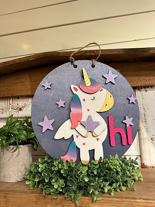 Decorative sign with a unicorn and 'hi' text on a wooden surface with plants.