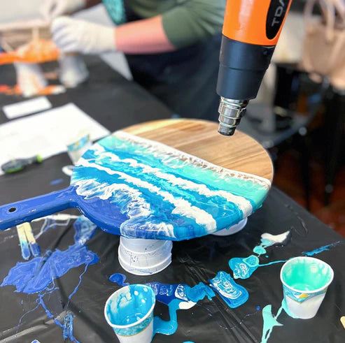 A hand-held heat gun is being used to apply heat to a round wooden board covered with blue and white paint in a swirling wave pattern. The setup is on a black table cover with paint cups, brushes, and paint splatters visible, and a person wearing gloves is blurred in the background.