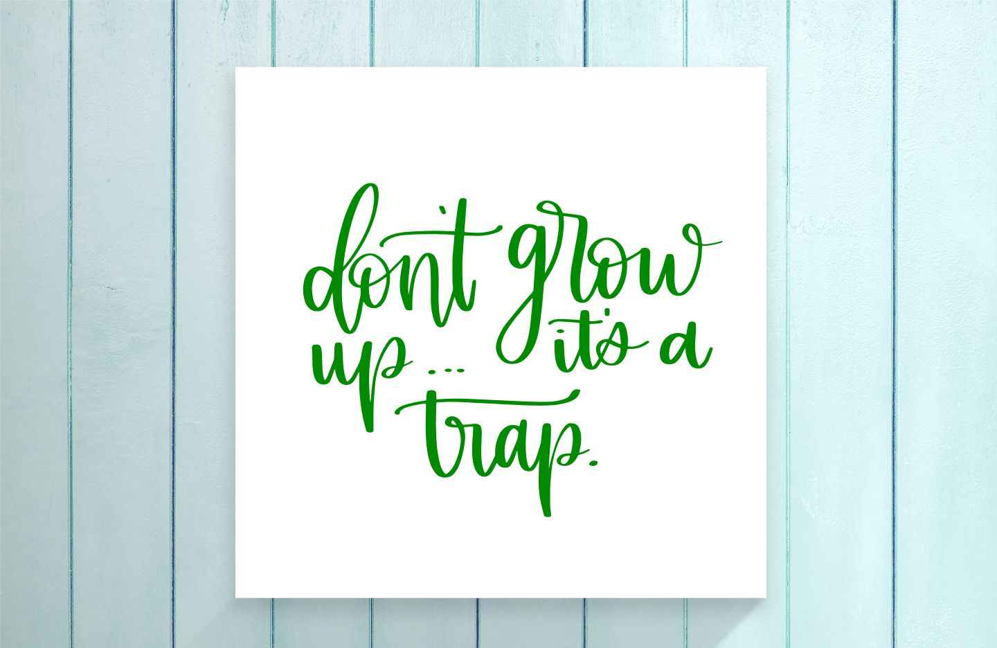 White card with green text 'don't grow up... it's a trap.' on a light blue wooden background