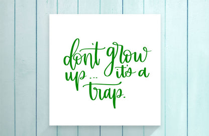 White card with green text 'don't grow up... it's a trap.' on a light blue wooden background