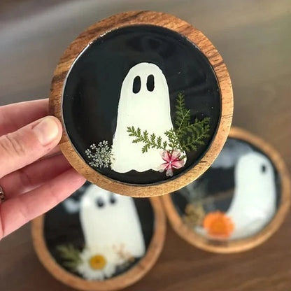 A hand holds a round wooden coaster featuring a black background with a white ghost design, green pressed leaves, and a small pink flower. Two more coasters are blurred in the background on a wooden surface.
