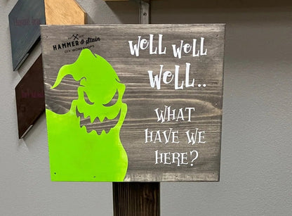 A rustic wooden sign on a dark wood stand with a painted green ghost and the text 'Well Well Well.