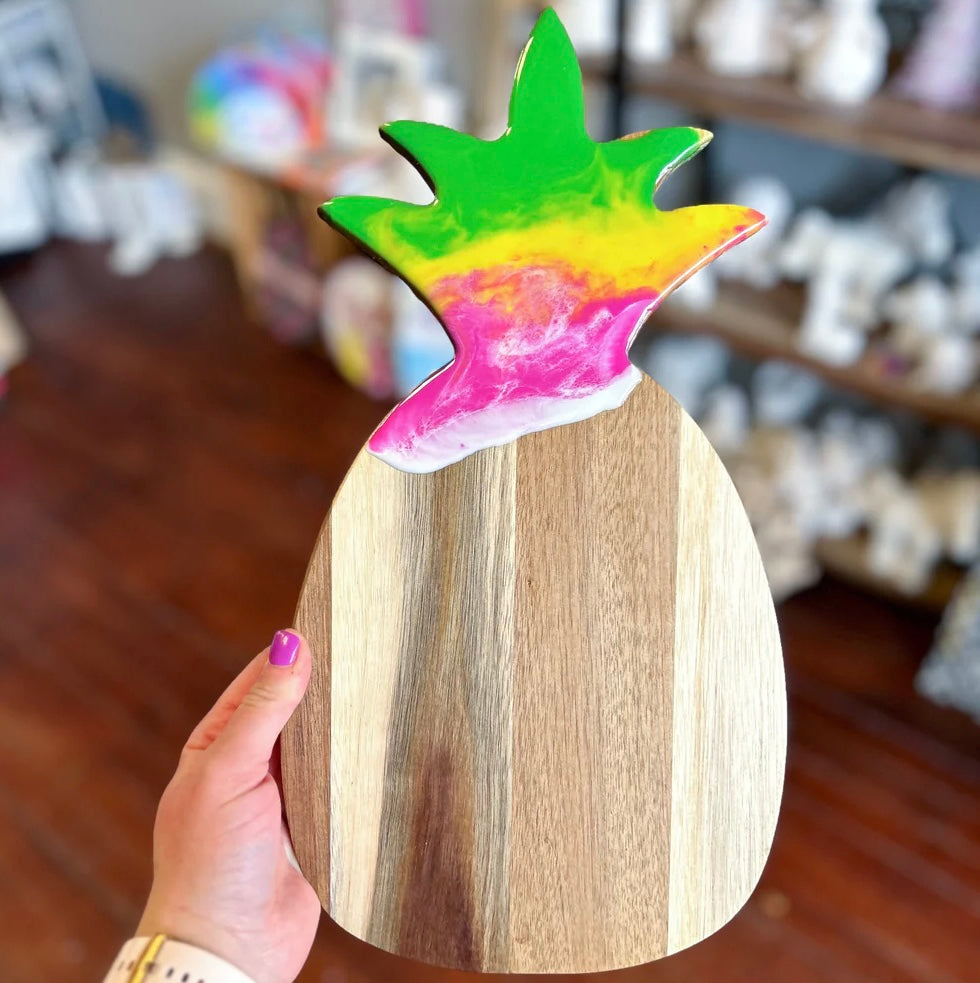 A hand with purple nail polish holds a wooden pineapple-shaped serving board or tray. The top of the pineapple is painted in bright green, yellow, pink, and white tie-dye style stripes, while the body is natural light brown wood.
