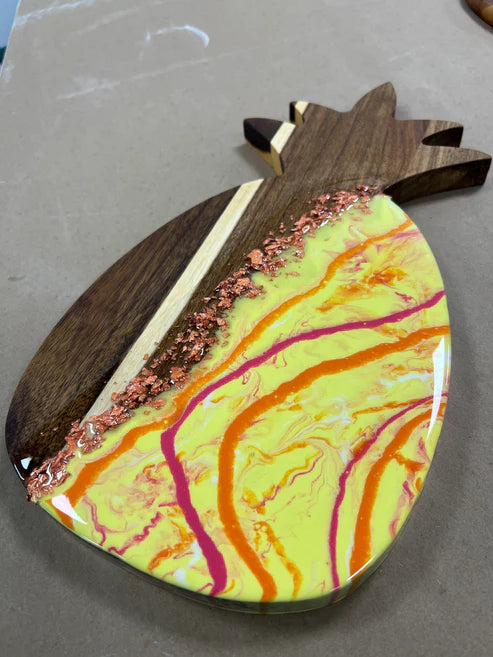 A pineapple-shaped wooden serving board or tray with a glossy yellow and orange marbled resin design, accented by pink swirls, rose gold glitter, and a dark wood handle.