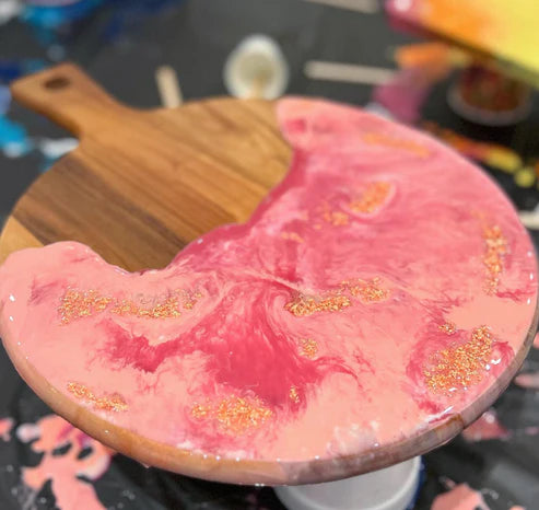 A round wooden serving board or tray with a handle, partially covered by a glossy pink and coral resin top featuring swirls and scattered gold glitter, photographed close up on a dark surface.