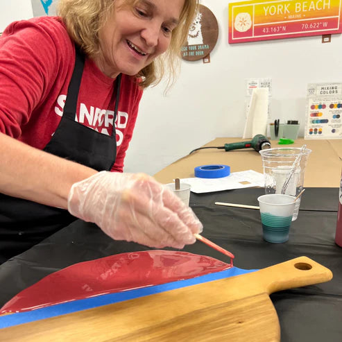 A woman wearing a red shirt, black apron, and clear plastic glove is painting a wooden serving board with red and blue paint in a workshop setting.