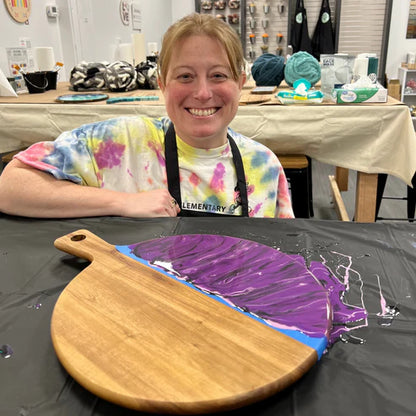 A smiling woman wearing a tie-dye shirt and black apron sits behind a craft table with a wooden paddle-shaped board covered in purple, blue, and white paint. The background shows a pottery or craft studio with tables, yarn balls, and ceramic items.
