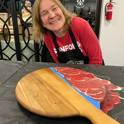 A smiling woman wearing a red shirt and black apron leans over a table with a large wooden paddle holding a painted red abstract design on a black surface.