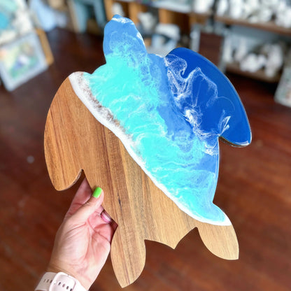 A hand holds a whale-shaped wooden plaque or tray with a layered resin ocean design in shades of blue, turquoise, and white, set against a blurred indoor background.