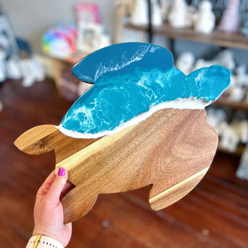 A hand holds a whale-shaped wooden plaque or tray with a layered resin ocean design in shades of blue, turquoise, and white, set against a blurred indoor background.