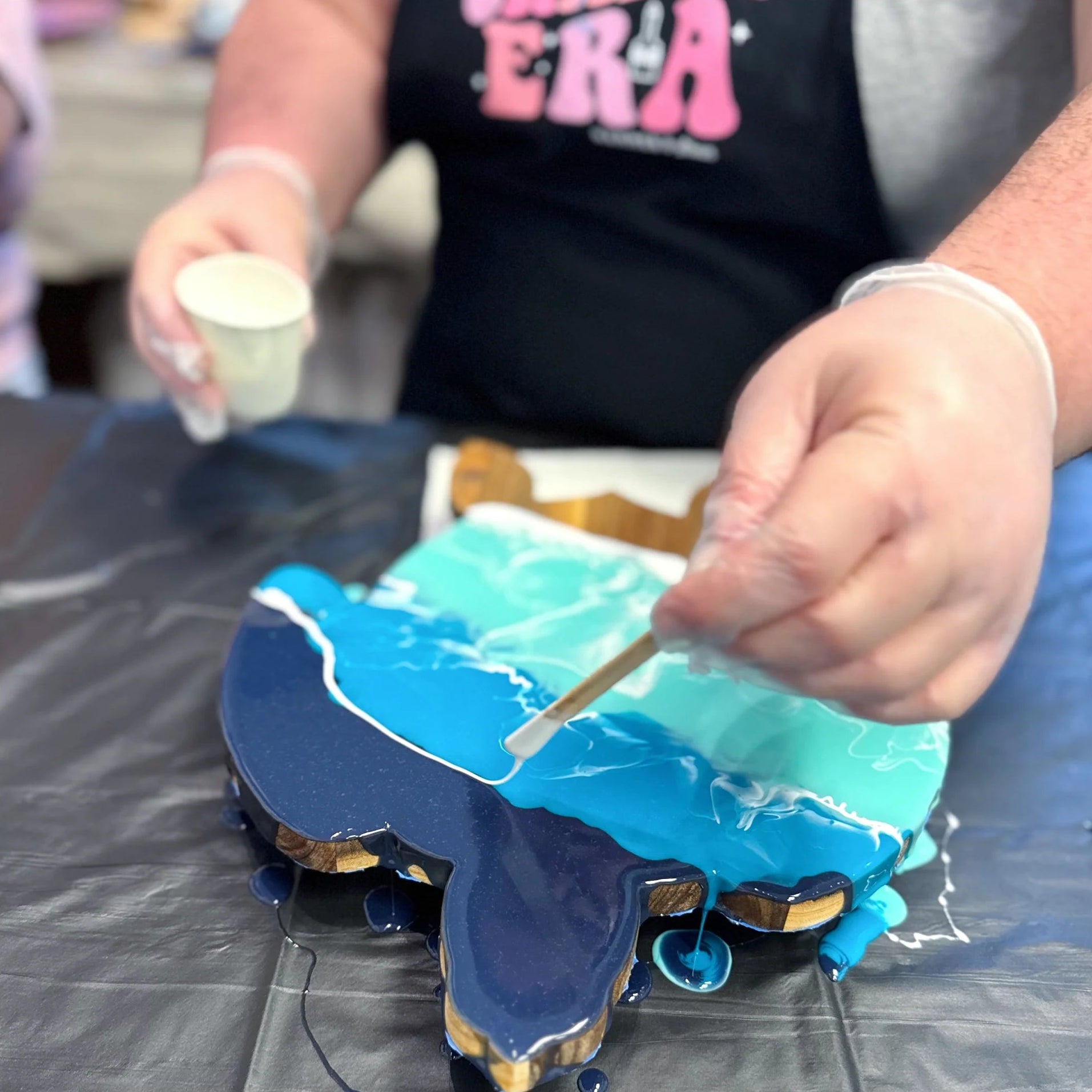 A close-up of a person wearing a black apron and clear gloves applying light blue icing to a shaped cake or decoration on a dark surface, with a small cup in the other hand.