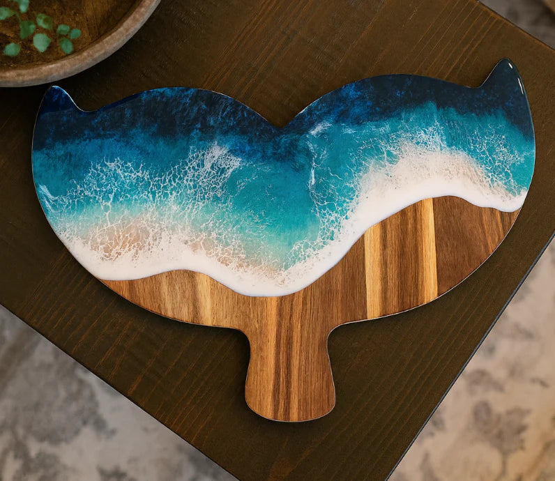A decorative serving board shaped like a whale or sea creature, featuring a printed ocean wave design in blue, teal, and white foam over a natural wood base, shown on a dark wooden table.